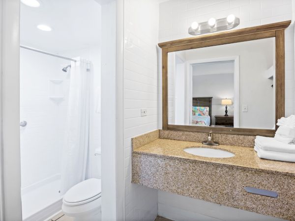 A modern bathroom with a shower, toilet, large mirror, sink, and towels on a granite countertop, featuring bright lighting.