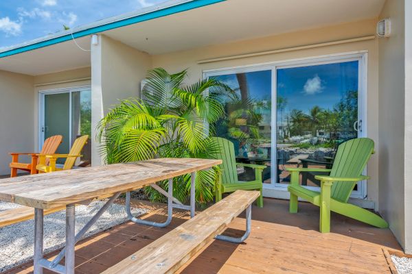 A patio with a wooden picnic table, colorful chairs, lush green plants, and sliding glass doors reflecting a sunny sky view.