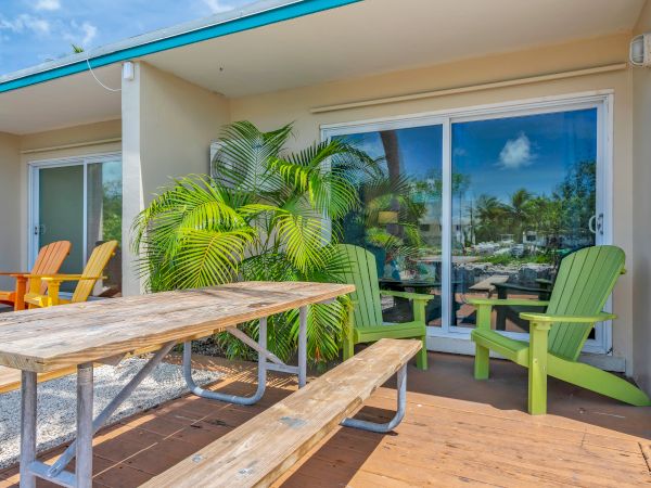 A patio with a wooden picnic table, colorful chairs, lush green plants, and sliding glass doors reflecting a sunny sky view.