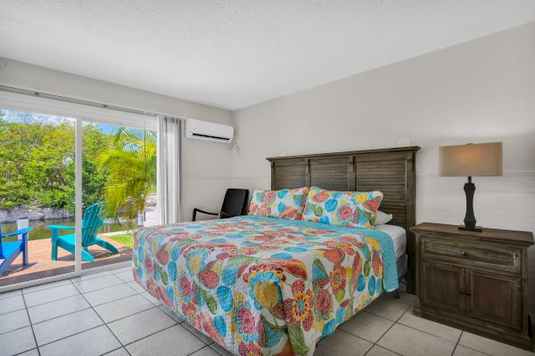 A bedroom with a colorful bedspread, a lamp on a nightstand, and a sliding door opening to a patio with chairs.