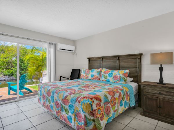A bedroom with a colorful bedspread, a lamp on a nightstand, and a sliding door opening to a patio with chairs.