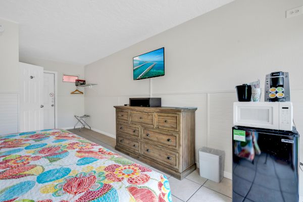 A hotel room with a colorful bed, dresser, wall-mounted TV, microwave, mini-fridge, and coffee maker in a bright and simple setup.