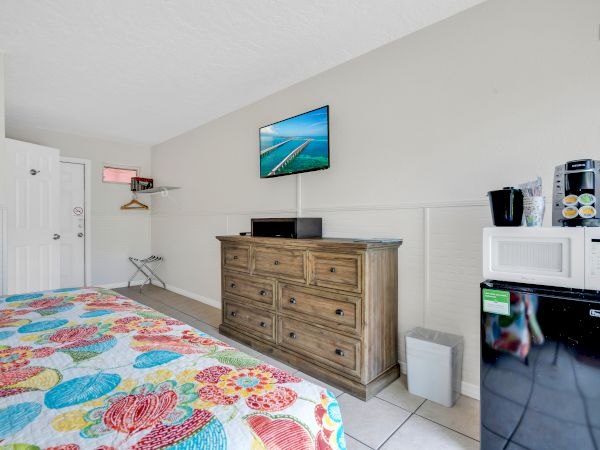 A hotel room with a colorful bed, dresser, wall-mounted TV, microwave, mini-fridge, and coffee maker in a bright and simple setup.