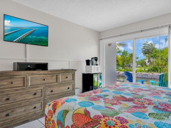 A colorful bedroom features a patterned bedspread, dresser, TV, and coffee maker; large windows reveal a pool and lush outdoor view.