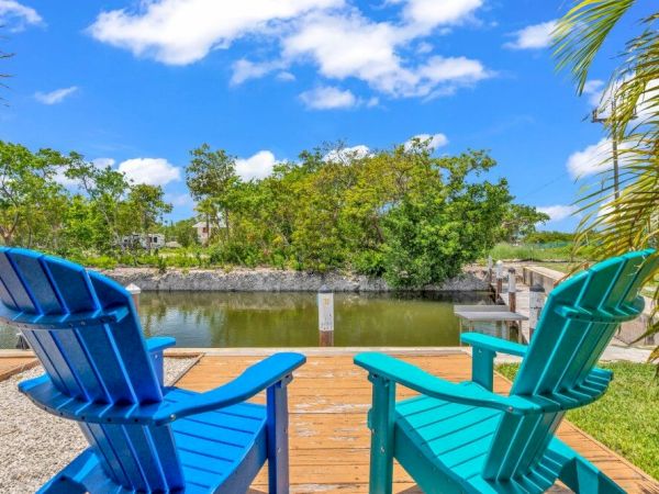 Two colorful chairs on a wooden deck overlook a tranquil canal surrounded by greenery, under a bright blue sky with fluffy clouds.