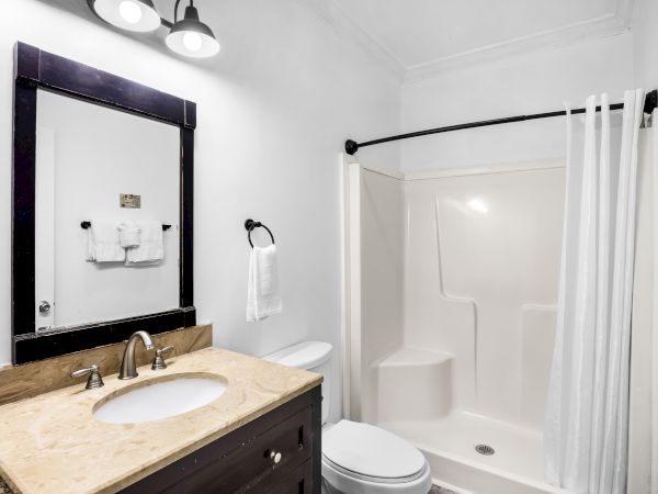 The image shows a bathroom with a sink, mirror, toilet, shower, and white towels. The decor is simple and modern.