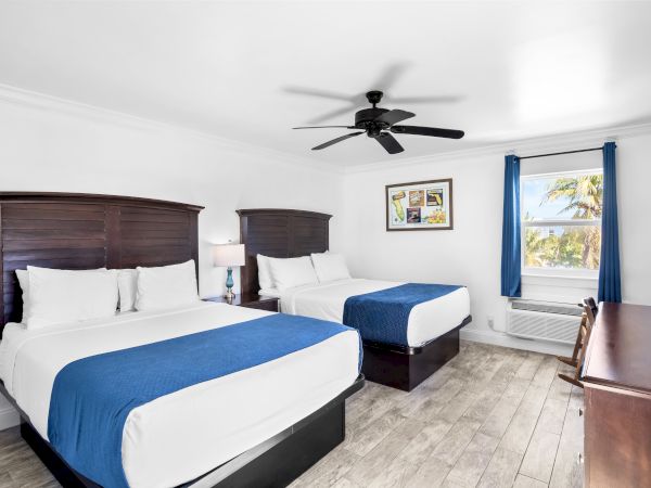 A hotel room features two beds with blue accents, a ceiling fan, a lamp, desk, and a window with blue curtains offering a tropical view.