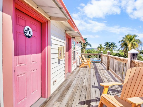 A wooden deck with pink doors numbered 307, wooden chairs, palm trees, and a bright sky create a relaxing tropical setting.