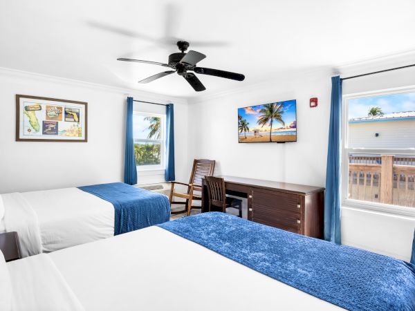 A bright hotel room features two beds, blue accents, a TV with a beach scene, a desk, chair, ceiling fan, and picturesque windows.