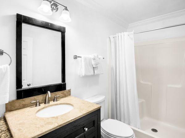 A bathroom with a marble countertop sink, mirror, towel racks, toilet, and a shower with a curtain under bright lighting.