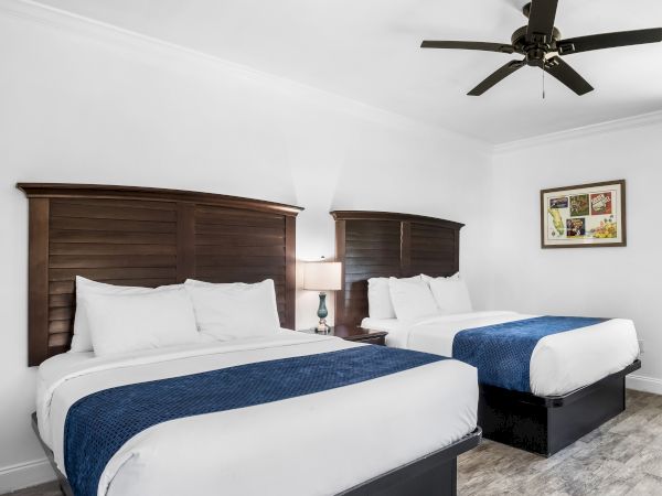 The image shows a hotel room with two beds, blue runners, wooden headboards, a lamp, artwork, and a ceiling fan.