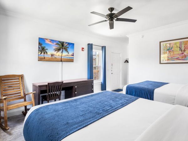 A hotel room with two beds, a rocking chair, desk, wall art, TV displaying a beach scene, and a ceiling fan is shown in the image.