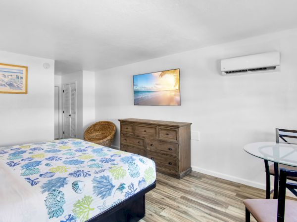The image shows a bedroom with a bed, dresser, air conditioner, wall art, wicker chair, and a small glass table with chairs.