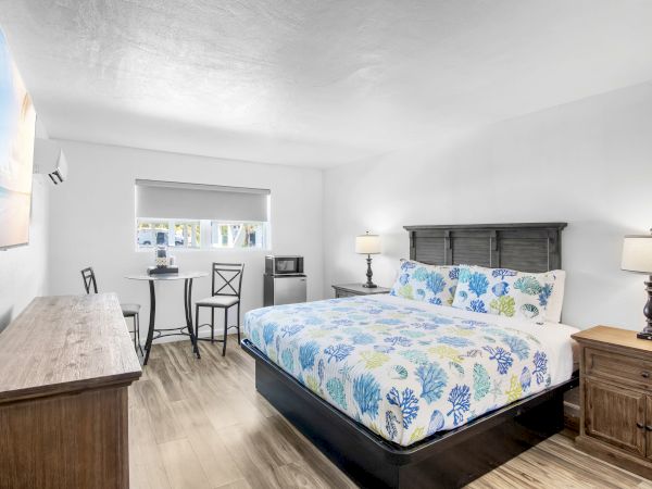The image shows a neatly arranged bedroom with a floral bed, a small table, chairs, a window, and lamps on wooden nightstands.