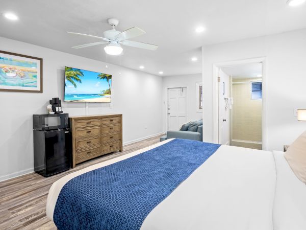 The image shows a cozy room with a bed, TV, dresser, microwave, fridge, and an open bathroom door. A ceiling fan is above.