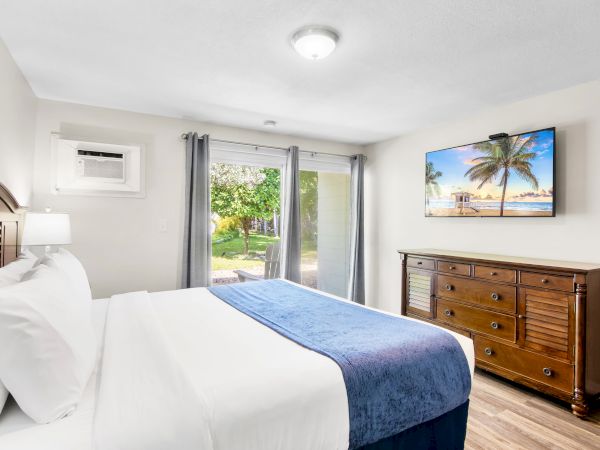 A bedroom with a neatly made bed, a wooden dresser, a wall-mounted TV, and a view of a garden through sliding glass doors.