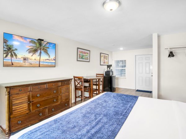 A hotel room featuring a bed with a blue runner, a wooden dresser, a small table with chairs, and a tropical beach painting on the wall.