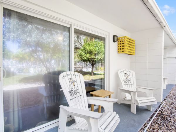 Two white Adirondack chairs and a small table are on a patio with a sliding glass door and a view of trees and a lawn outside.