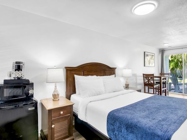A hotel room with a bed, two lamps, a small fridge, a microwave, and a table with chairs by the window ending the sentence.
