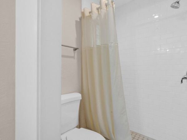 The image shows a bathroom with a toilet, a shower area with a white curtain, and white tiled walls.