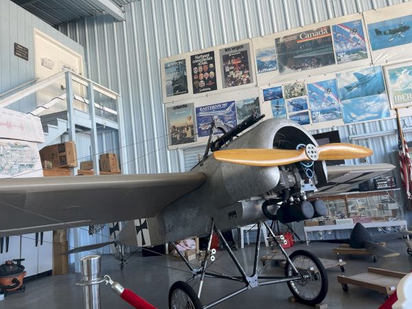 The image shows a vintage aircraft displayed in a museum, surrounded by aviation-themed posters on a corrugated metal wall.