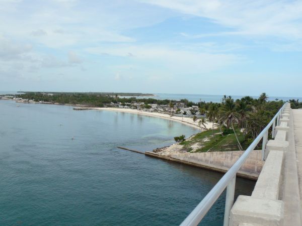 A coastal view from a bridge with clear blue water, lush green trees, and a small settlement by the shoreline under a partly cloudy sky.