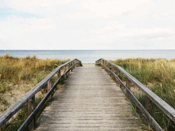 A wooden boardwalk leads through tall grass towards a beach with a calm sea and cloudy sky, creating a serene coastal view.