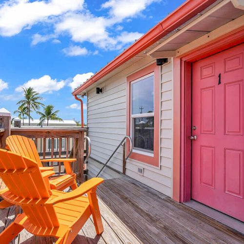 A porch with two orange chairs, a pink door, wooden railings, and a view of palm trees under a blue sky with clouds.