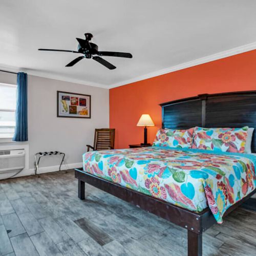 A bedroom with colorful bedding, two lamps, a ceiling fan, and wood flooring. An orange accent wall and a window are visible.