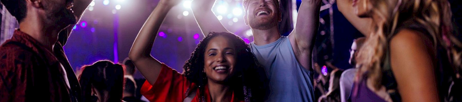 A group of people enjoying a concert, dancing and smiling, with stage lights and a lively atmosphere in the background.
