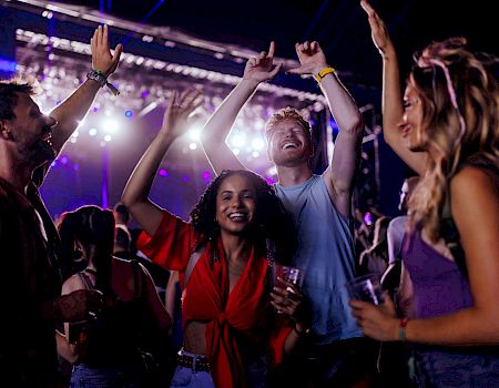A group of people enjoying a concert, dancing and smiling, with stage lights and a lively atmosphere in the background.