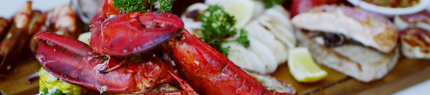 A seafood platter featuring lobster, baked potato, corn on the cob, and various sauces on a wooden board garnished with herbs.