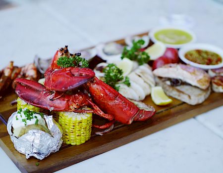 A seafood platter featuring lobster, baked potato, corn on the cob, and various sauces on a wooden board garnished with herbs.