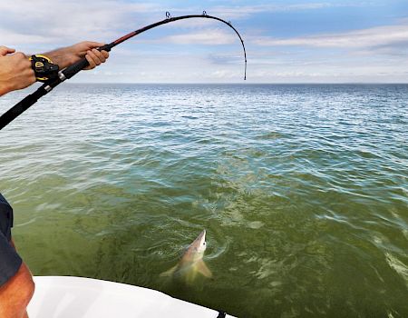 A person is fishing from a boat in the ocean, reeling in a fish that's partially visible in the water near the surface.