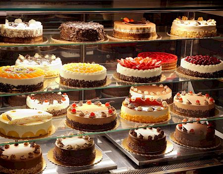 A variety of decorated cakes displayed in a glass case, each with unique toppings and designs, stacked on multiple shelves in a bakery.