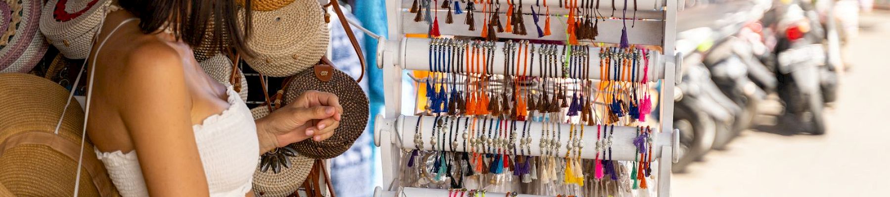 A person browsing a market stall displaying colorful bracelets and hats. Scooters are lined up along the street in the background.