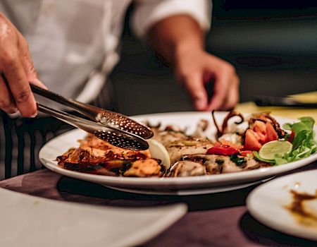 A chef is using tongs to arrange grilled seafood and vegetables on a plate, which includes fish, lime, and greens, on a dining table.