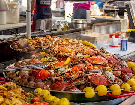 The image shows a food stall with a large platter of seafood, including crabs and shrimp, surrounded by lemons and cooked potatoes.