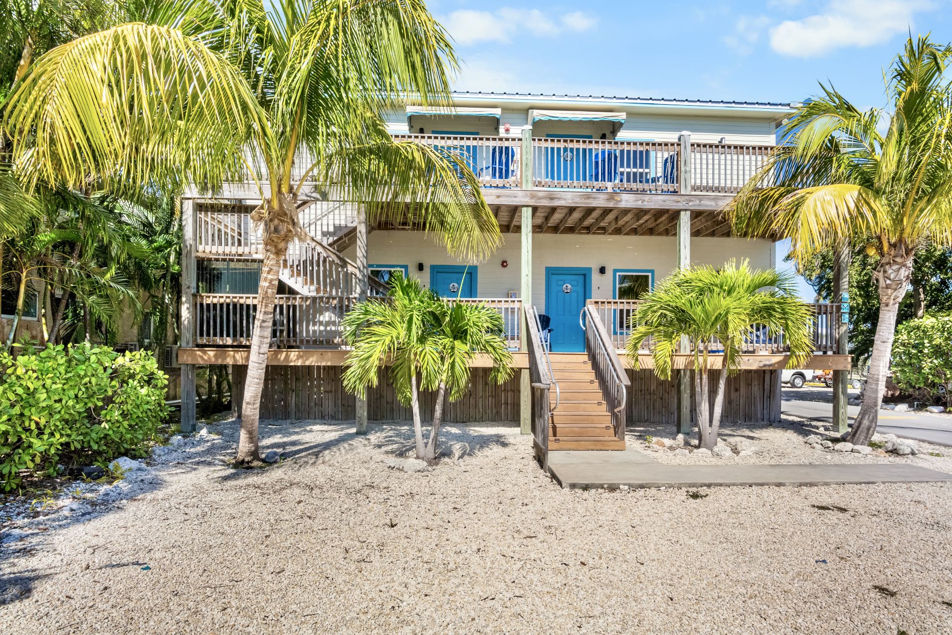 A tropical beach house with blue doors, surrounded by palm trees and bright sunshine.