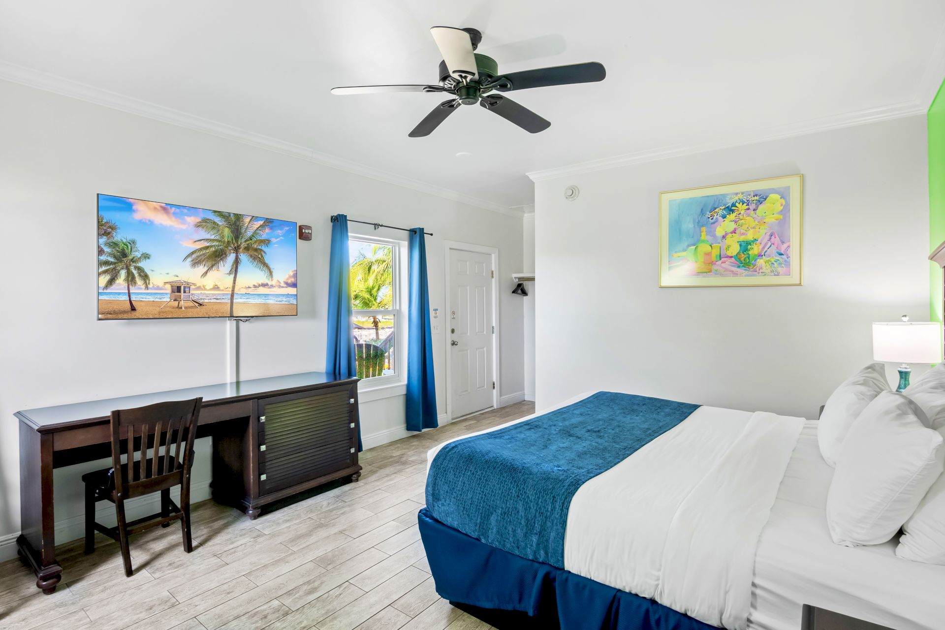 A clean, modern hotel room with a bed, desk, TV showing a beach scene, colorful artwork, and a window with blue curtains.