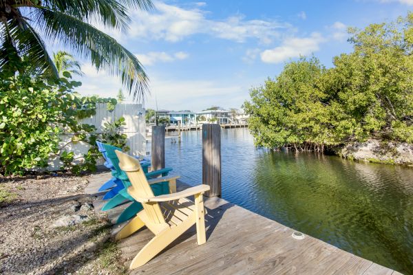 Wooden dock with colorful chairs overlooks a calm canal, surrounded by lush greenery and houses under a clear blue sky.