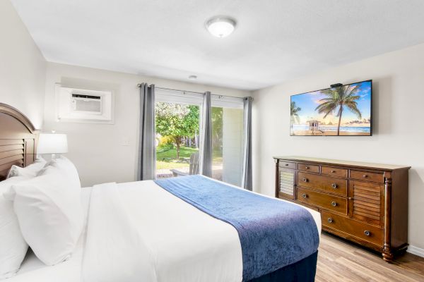 A cozy bedroom features a bed with white linens, a blue throw, wooden dresser, wall-mounted TV, window view of greenery, and an air conditioner.