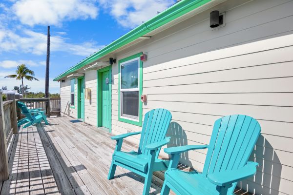 A wooden deck with turquoise Adirondack chairs in front of a building with green-trimmed doors and windows under a sunny sky.