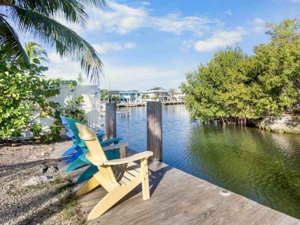 Wooden dock with colorful chairs overlooks a calm canal, surrounded by lush greenery and houses under a clear blue sky.