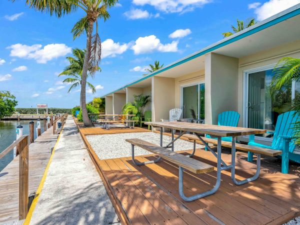 A sunny waterfront scene with a row of modern cottages, palm trees, blue sky, and a wooden deck featuring a picnic table and colorful chairs along the canal.