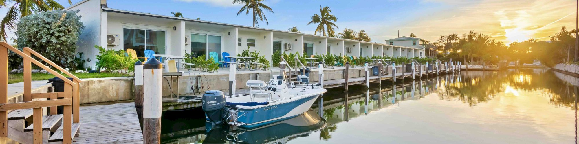 A peaceful waterfront scene with boats docked along a calm canal, white waterfront homes, palm trees, and a golden sunset reflecting on the water.