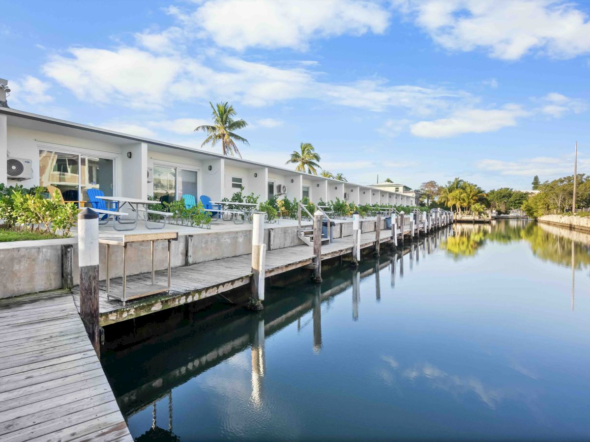 A row of white cottages along a calm canal with wooden docks, clear blue sky, and palm trees reflected in the water.