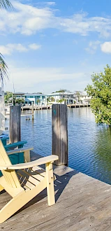 Two wooden adirondack chairs sit on a weathered dock by calm water, with palm trees and mangroves lining a sunny, tropical shoreline.