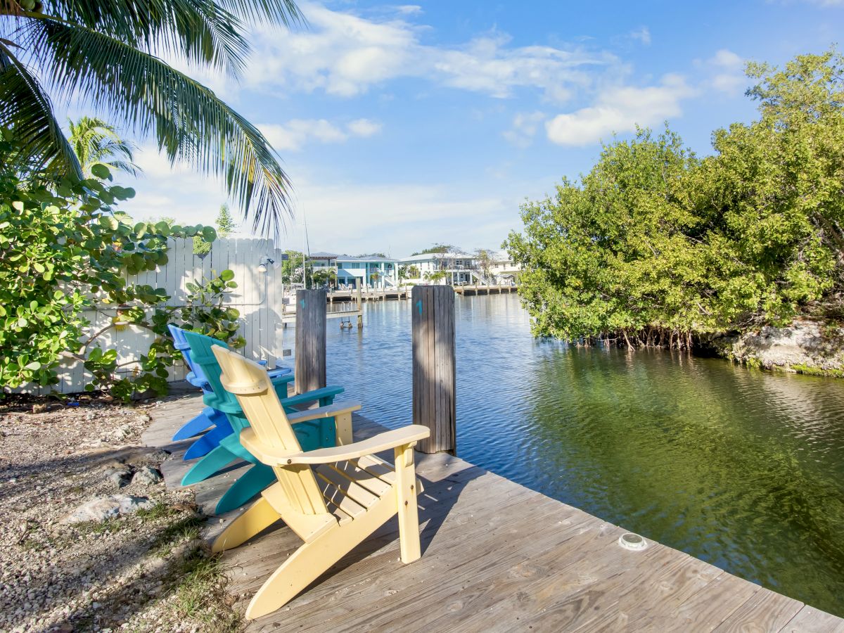 Two wooden Adirondack chairs sit on a weathered dock by calm water, with tropical trees and a bright sky in the background.