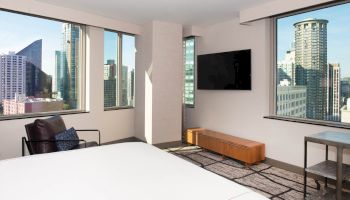 A modern hotel room with large windows showing a city view, featuring a bed, chair, TV, and minimalist decor.